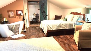 Ein gemütliches Schlafzimmer im Dachgeschoss mit einem großen Bett, einem kleineren Bett und einem Sessel. Der Boden ist Holzdielen und es gibt eine Deckenleuchte.
