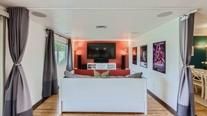 A modern living room with a white sofa, a flat-screen TV mounted on a red brick wall, and a dining area with a table and chairs. - Arizona Daydream + Private Pool + EV Charger (Chandler)