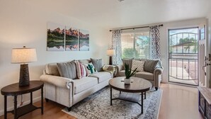 A living room with a beige sofa, two side tables with lamps, a coffee table, and a patterned rug. There are two large windows with patterned curtains and a view of a building outside. - Arizona Daydream + Private Pool + EV Charger (Chandler)