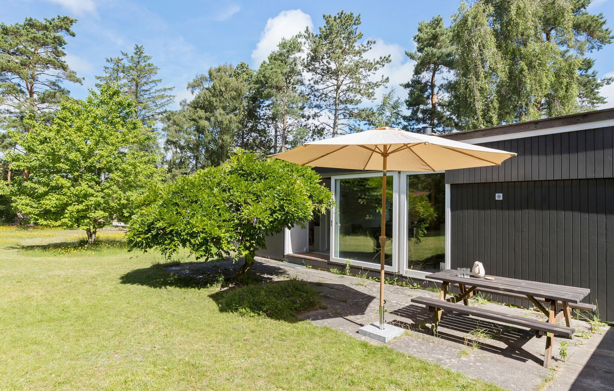 Un patio con una mesa y bancos de madera, un paraguas grande y una puerta de vidrio que da a un espacio exterior cubierto.