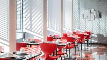 A modern restaurant with red chairs and tables, large windows with blinds, and a polished floor.
