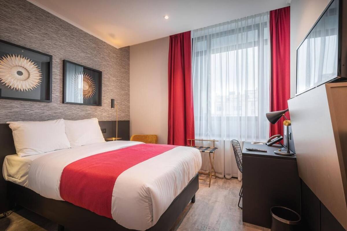 A modern hotel room with a large bed, a desk with a chair, a desk lamp, and a large window with red curtains.