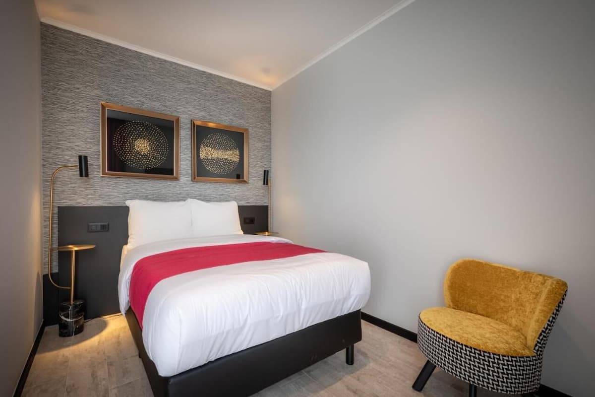 A modern hotel room with a large bed, two wall-mounted artworks, a yellow chair, and a small side table with a lamp.