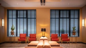 A hotel room with a large bed, two red chairs, a table, and large windows with blinds. - Hotel Jamingo (Antwerp)