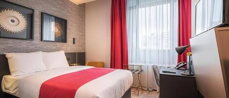 Quarto de hotel com uma cama grande, uma escrivaninha com luminária, uma cadeira e duas obras de arte emolduradas na parede.
