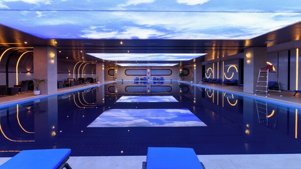 A modern indoor swimming pool with a clear view of the sky through a ceiling design, lounge chairs, and a ladder for pool access. - Rumanza by Pearl Continental (Multan)