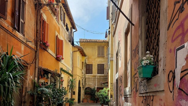 A narrow cobblestone alley flanked by buildings with balconies and potted plants. - 4bnb - Lungaretta Jacuzzi Apartment (Rome)