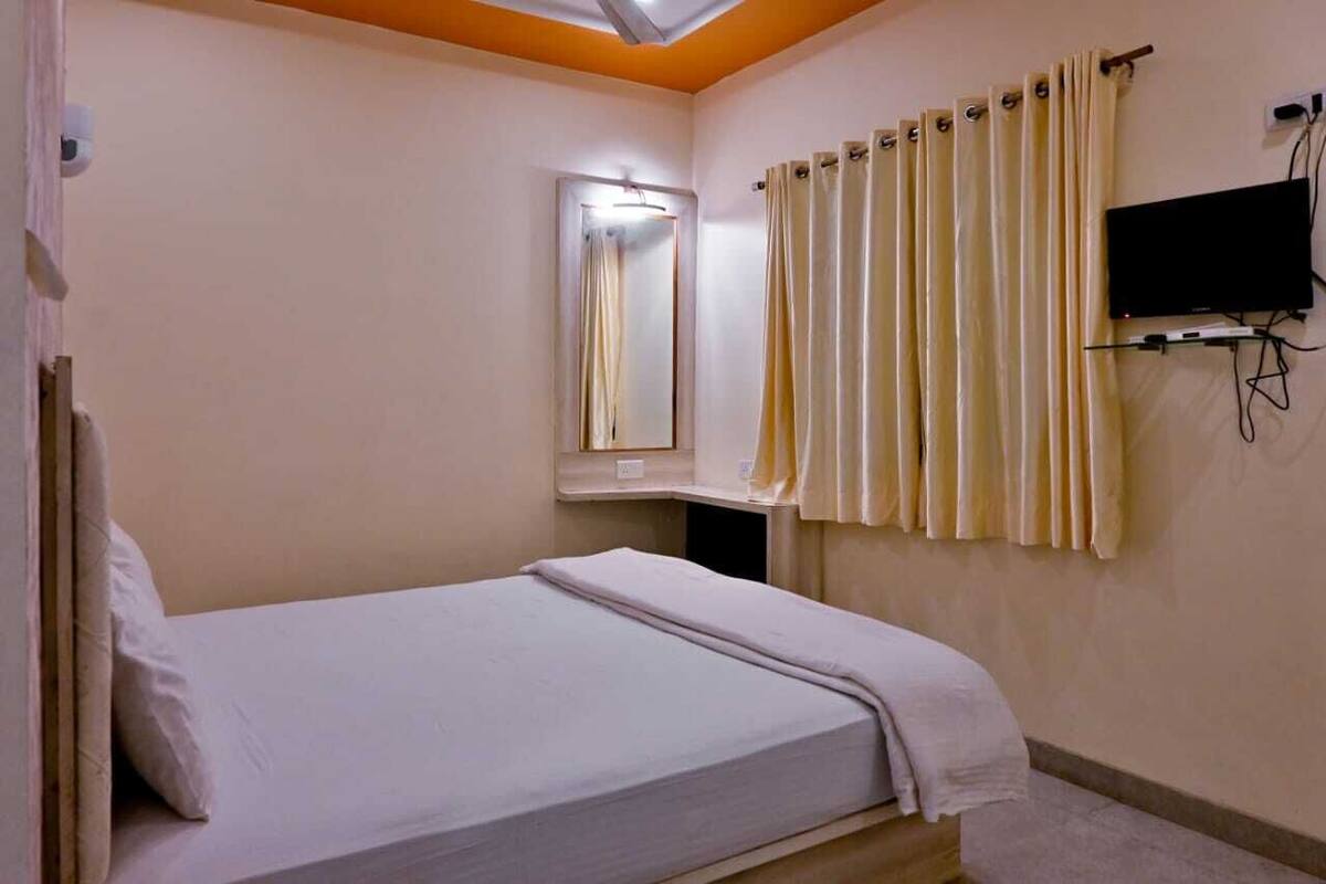 A bedroom with a bed, a mirror, a TV mounted on the wall, and a window with curtains.