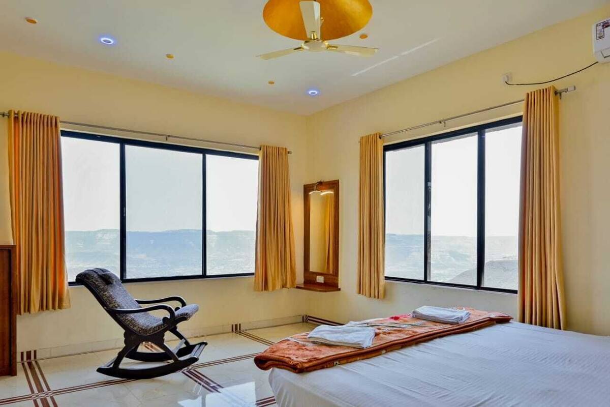 A hotel room with a bed, a rocking chair, a large window with curtains, and a ceiling fan.