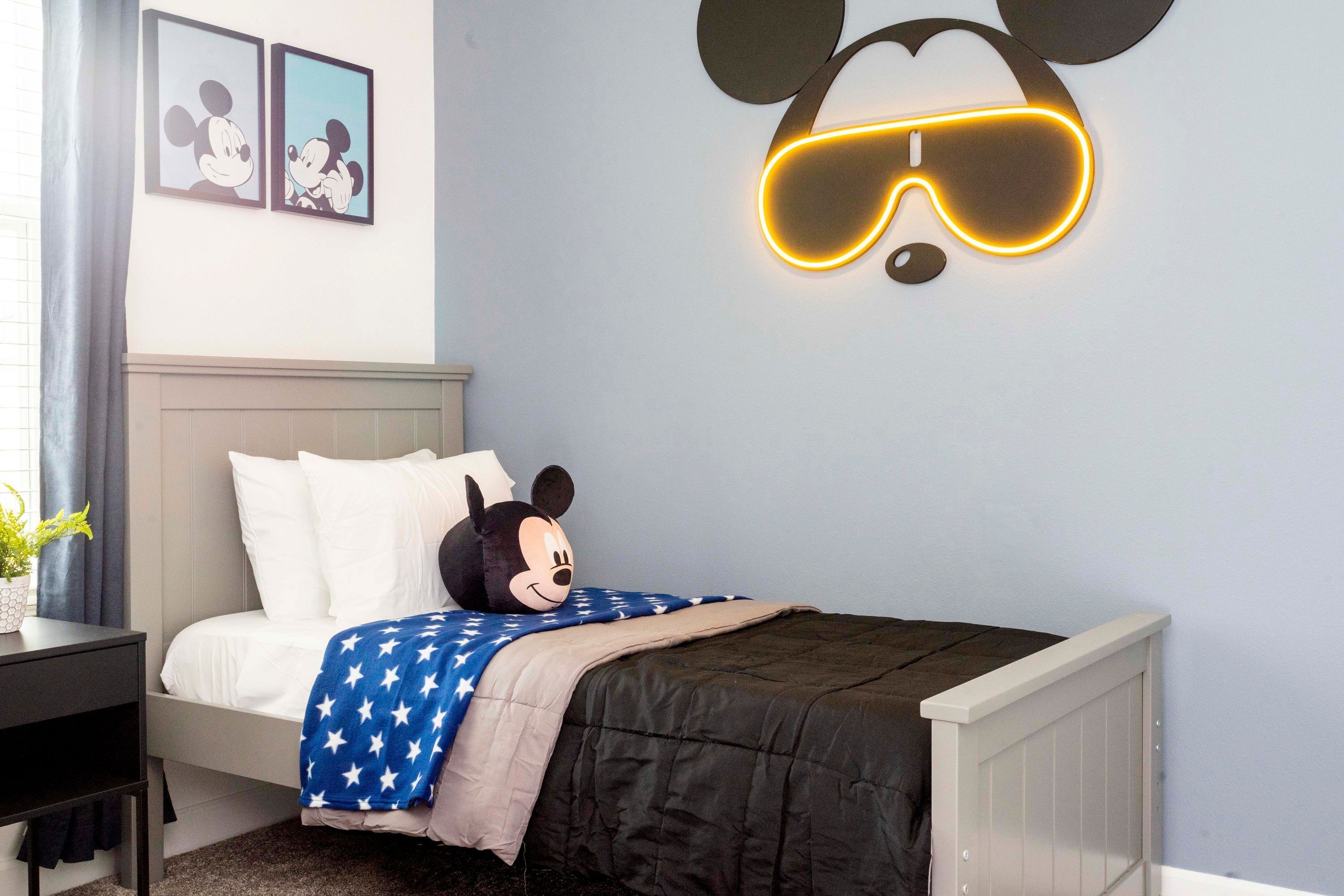 Un dormitorio con cabecera temática de Mickey Mouse, una cama con manta azul y una mesita de noche con una planta.