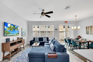 A modern living room with a sofa, coffee table, and TV stand. There is a dining area with a table and chairs.