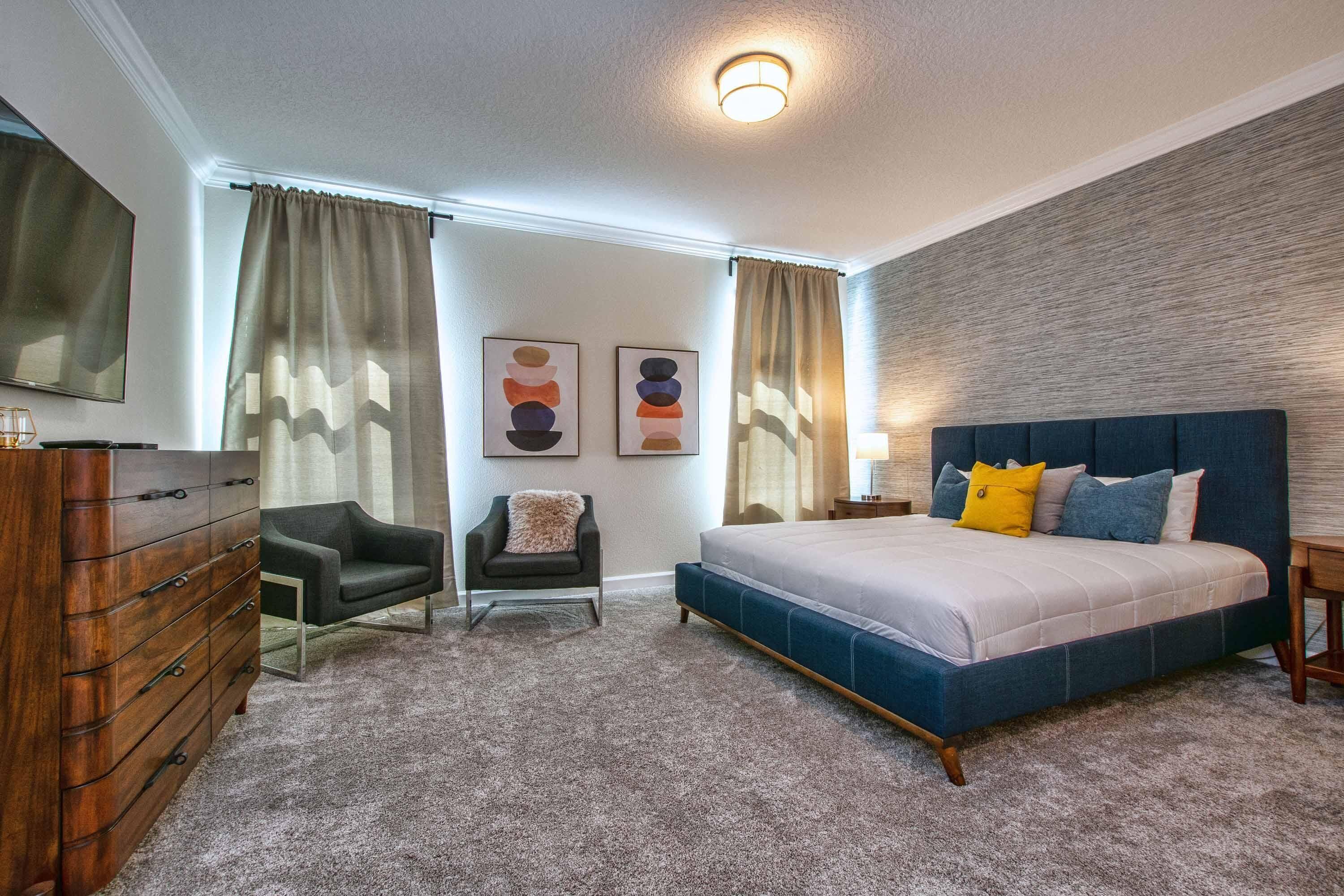 A bedroom with a bed, dresser, chair, and artwork on the wall.
