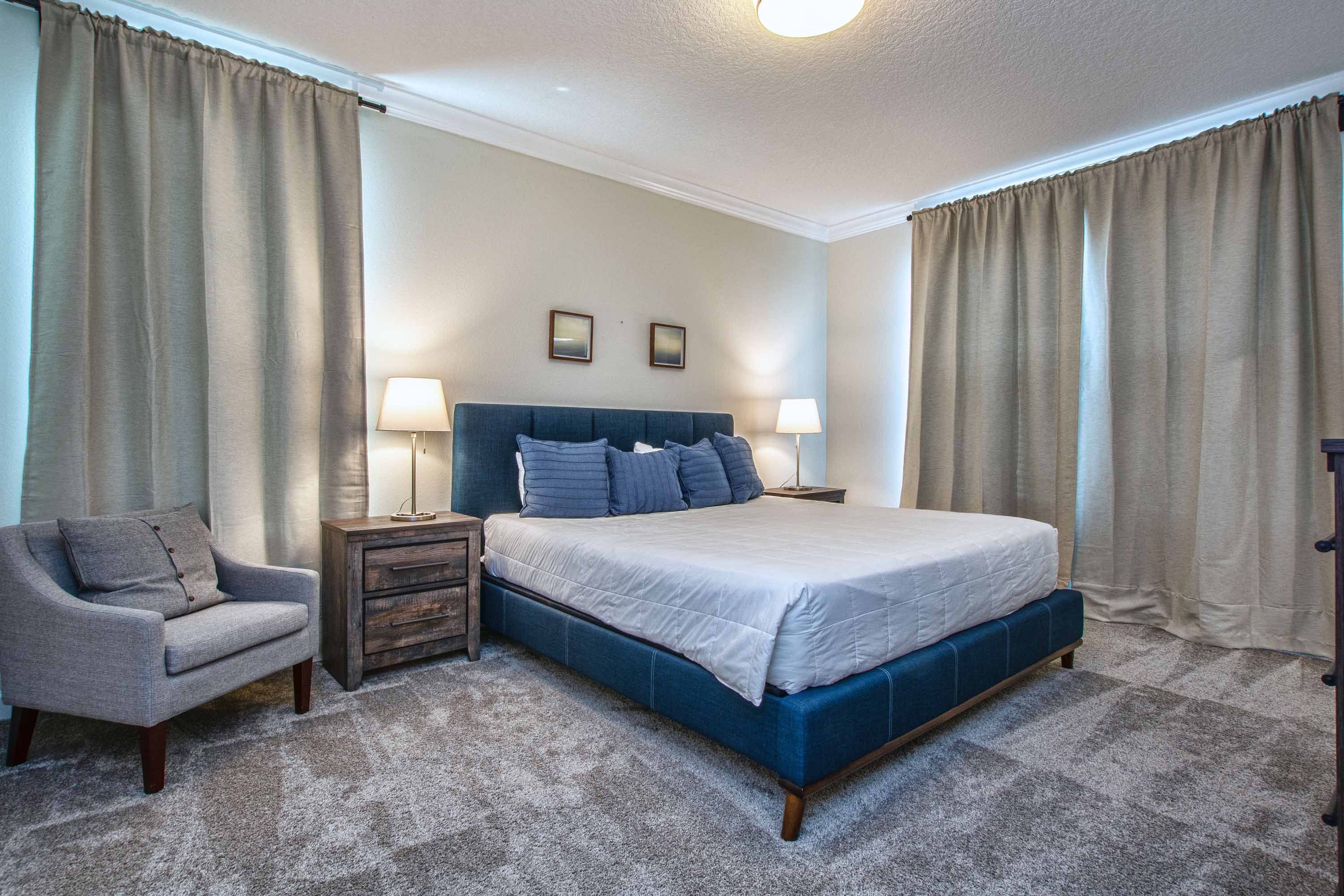 A bedroom with a large bed, two bedside tables with lamps, a chair, and beige curtains.