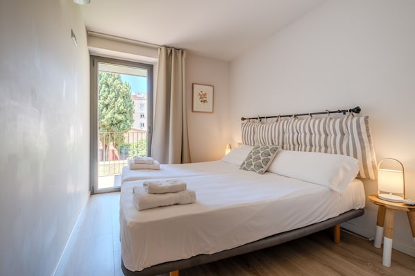 A bedroom with a bed, a bedside table with a lamp, a window with curtains, and a framed picture on the wall.