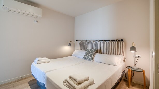 Een net opgemaakt bed met witte lakens en een opgevouwen handdoek, een bedlamp en een aan de muur bevestigde airconditioner.