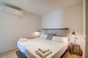 A neatly made bed with white linens and a folded towel, a bedside lamp, and a wall-mounted air conditioner.