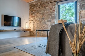 A modern living room with a stone wall, a flat-screen TV mounted on the wall, a wooden floor, and a large window providing natural light.