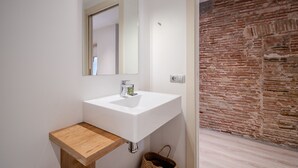 Een badkamer met een wit wastafel, houten opbergmeubel en een grote spiegel. Een bakstenen muur is op de achtergrond zichtbaar.