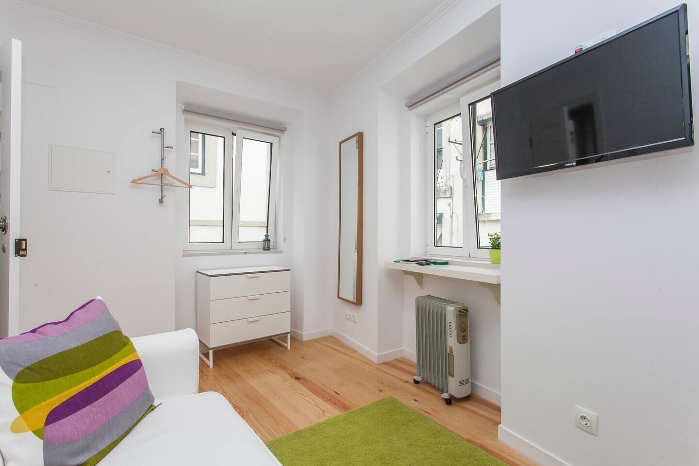A small, bright room with a white dresser, a bed with a colorful pillow, a radiator, and a TV mounted on the wall.