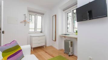 A small, bright room with a white dresser, a bed with a colorful pillow, a radiator, and a TV mounted on the wall.
