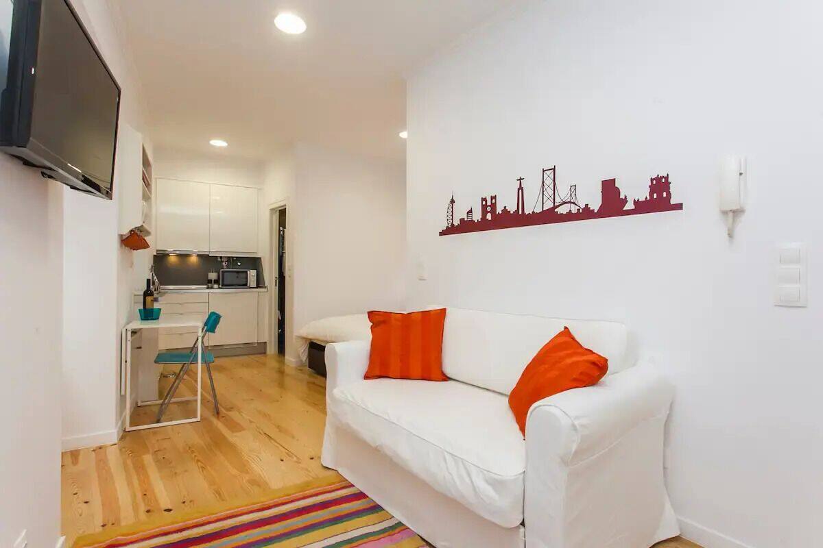 Un espace de vie compact avec un canapé blanc, des coussins orange, une décoration murale représentant un paysage urbain, un coin cuisine et un plancher en bois.
