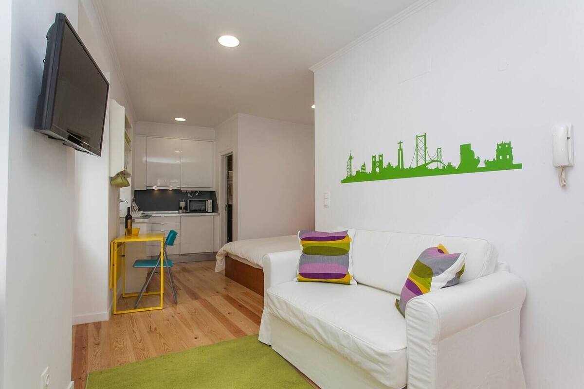 Un espace de vie compact comprenant un coin cuisine, un espace repas avec une table jaune et une chaise bleue, un canapé blanc agrémenté de coussins colorés et un décor mural représentant un paysage urbain verdoyant.