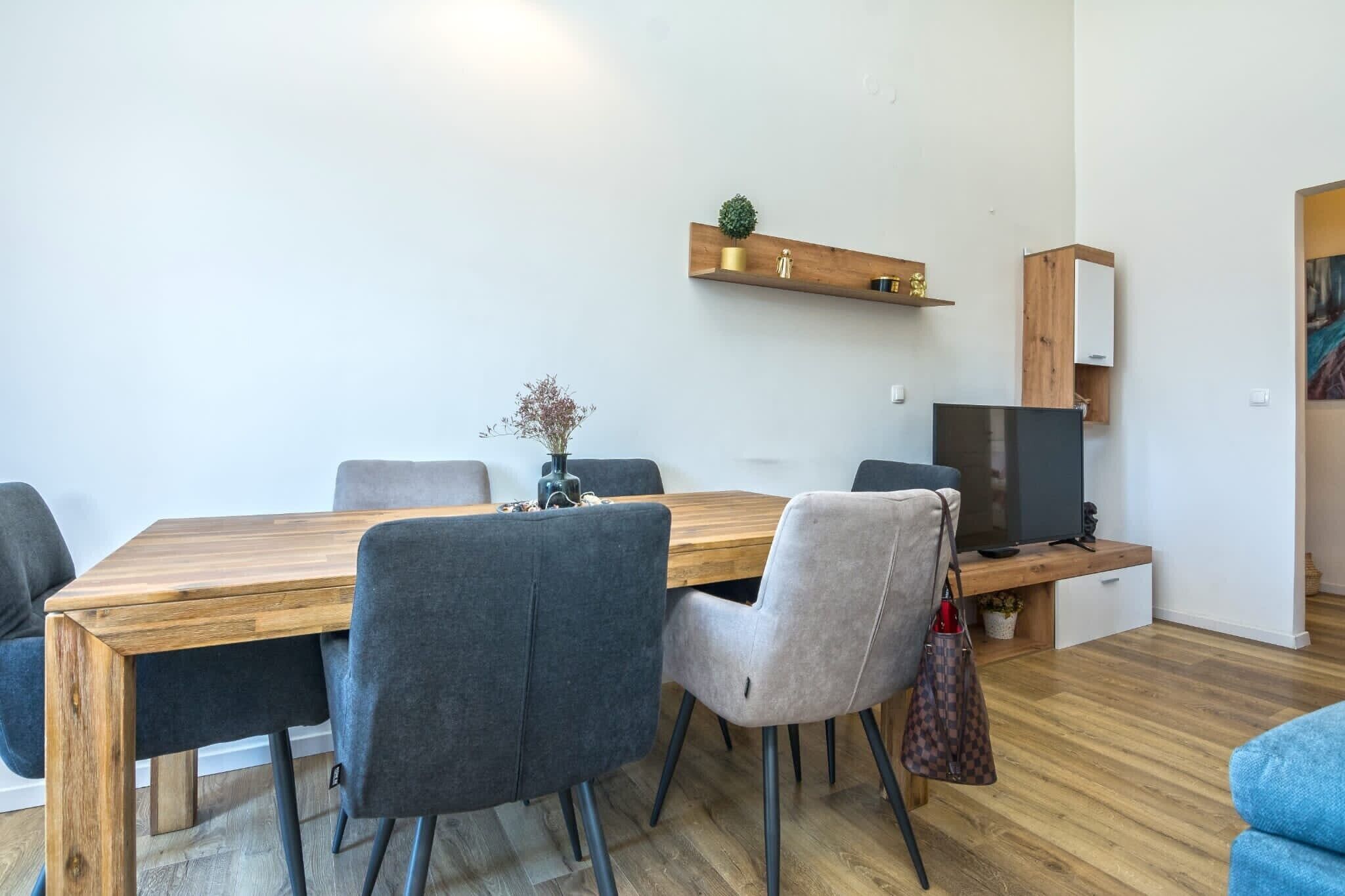 A modern dining area with a wooden table, grey upholstered chairs, a television on a wooden stand, and a small shelf with decorative items.