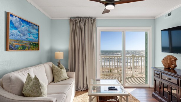 A living room with a beige sofa, a glass coffee table, a ceiling fan, a painting, a television, and a view of the beach through a sliding glass door. - Pup-Friendly! Direct Oceanfront! Shorely Goodness: PierView 105 Oceanfront Condo (Nags Head)