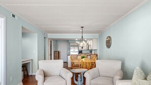 A living room with two armchairs, a small round table, and a sofa. There is a dining area with a wooden table and chairs in the background. - Pup-Friendly! Direct Oceanfront! Shorely Goodness: PierView 105 Oceanfront Condo (Nags Head)