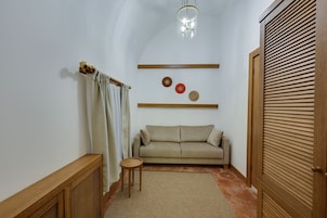 Um corredor estreito com um sofá bege, uma pequena mesa redonda e uma porta de madeira com ripas horizontais.