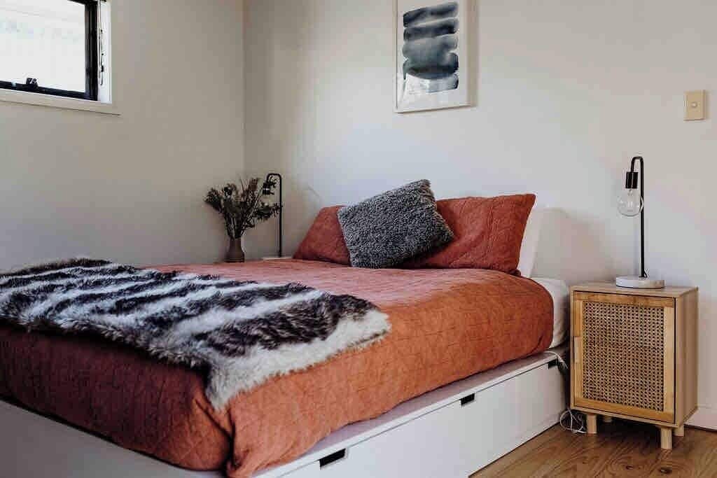 Un dormitorio con cama, mesita de noche con lámpara, una obra de arte en la pared y una ventana.