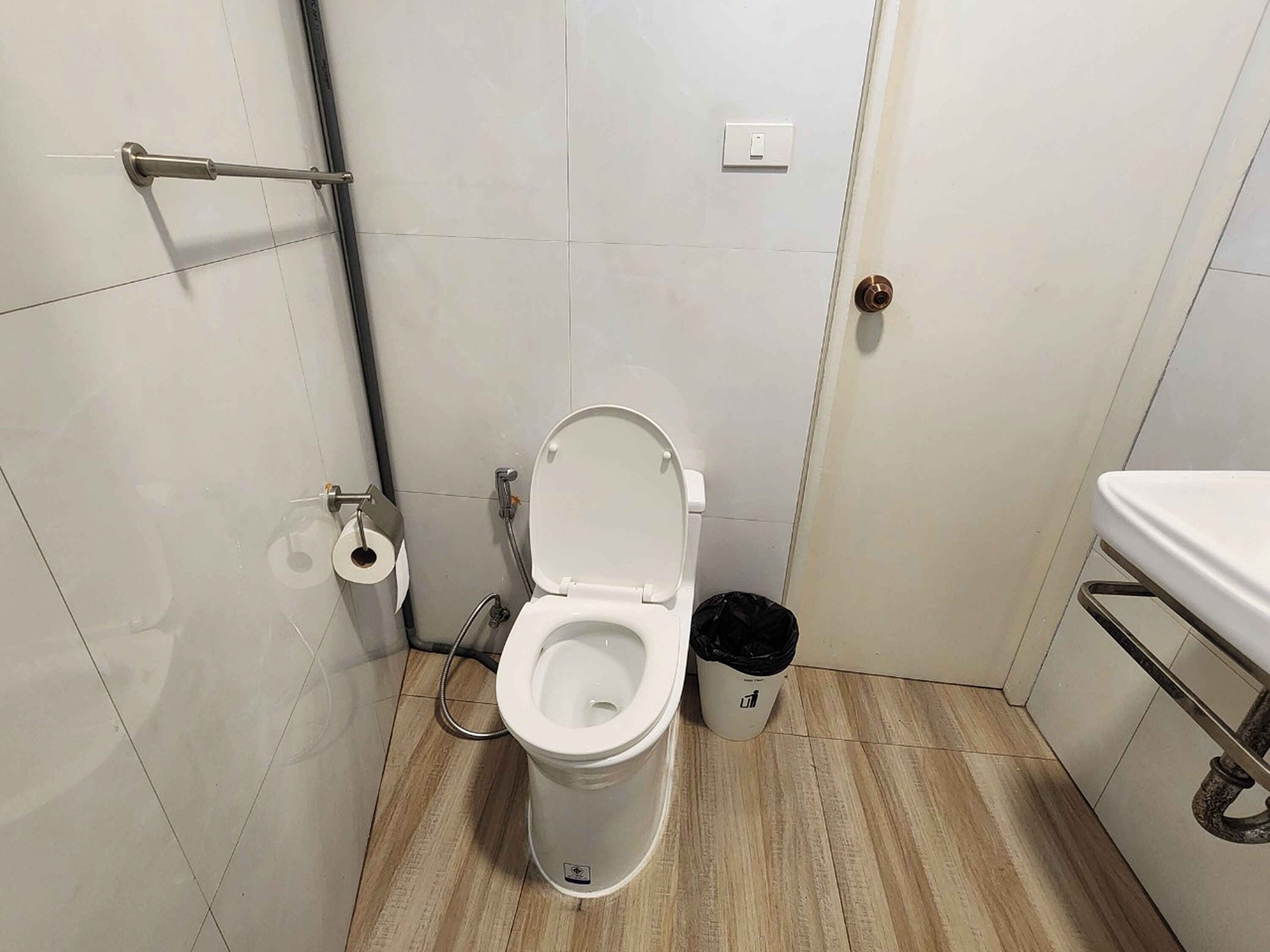 Een kleine, schone badkamer met toilet, prullenbak, toiletrolhouder en bedieningspaneel voor de douche.