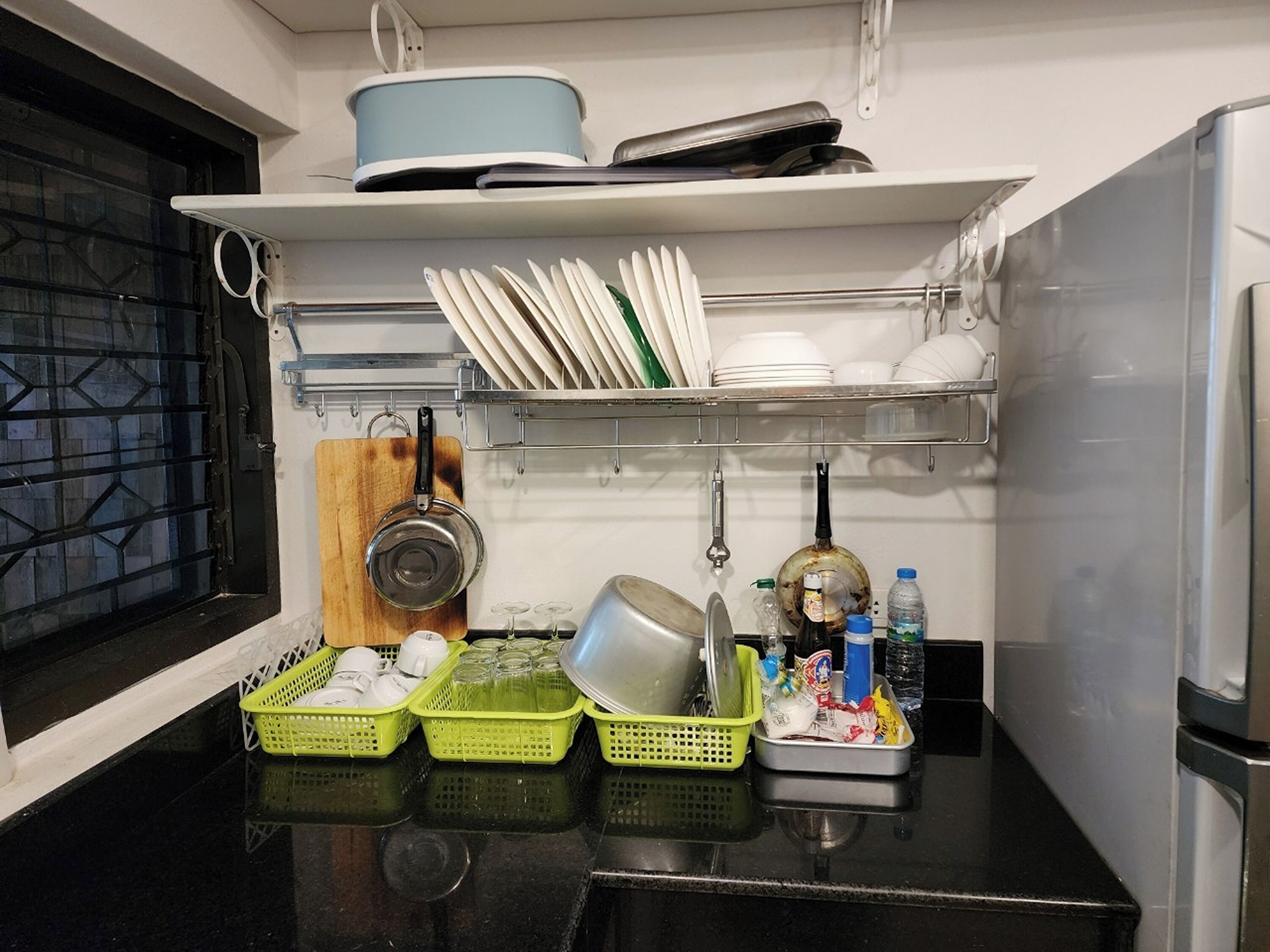 A kitchen with a black countertop, a window with a metal grill, and shelves holding dishes and kitchen utensils.