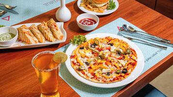 Uma mesa de jantar com diversos pratos, incluindo um hambúrguer, batatas fritas, uma pizza e uma salada. Há um copo de cerveja e uma rosa em um vaso.