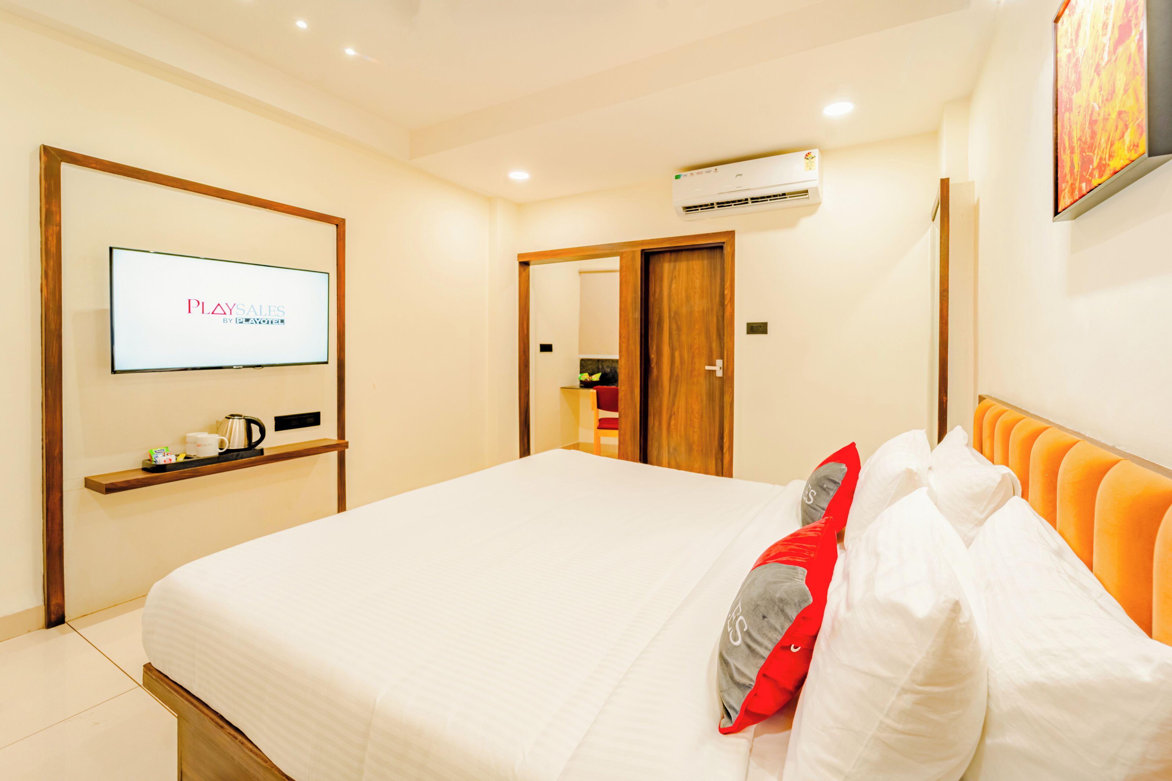 A hotel room with a bed, a TV mounted on the wall, a shelf with a coffee pot, and an air conditioning unit.
