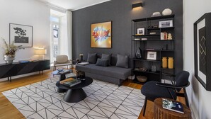 A modern living room with a grey sofa, black coffee table, and a geometric rug. There is a bookshelf with decorative items and a framed picture on the wall. - Exclusive City Hideaway (Lisbon)