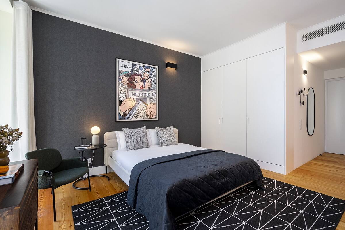 Ein modernes Schlafzimmer mit einem Bett, einem Nachttisch, einem Sessel, einem schwarz-weißen geometrischen Teppich und einem Bild an der Wand.
