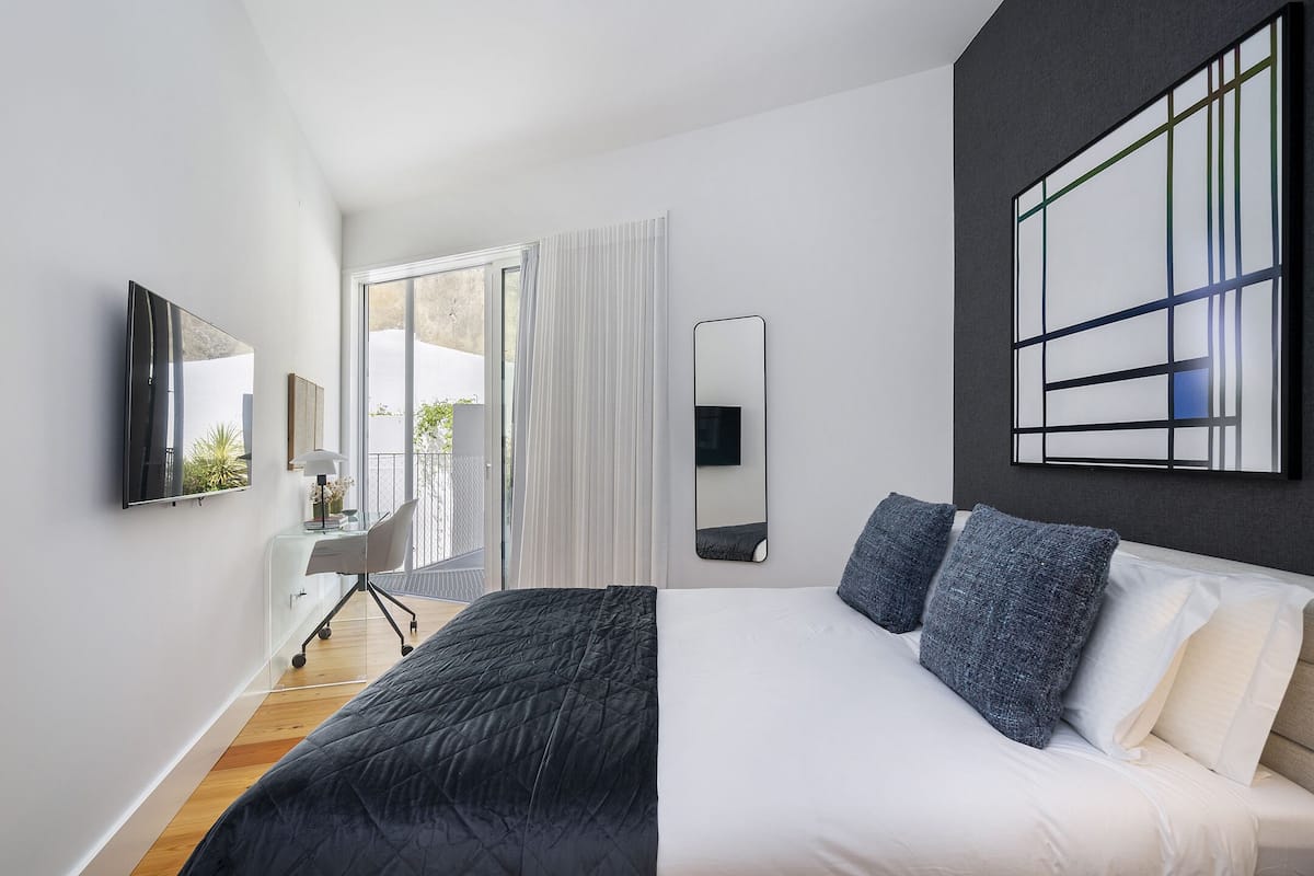Ein modernes Schlafzimmer mit einem großen Bett, einem Schreibtisch mit Stuhl, einem Fernseher und einem großen Fenster mit Vorhängen.
