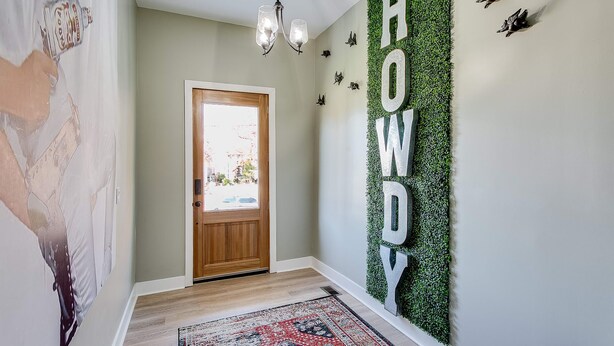 Un pasillo con una puerta de madera, una pared verde donde se lee la palabra "HOWDY" y un tapete estampado en el suelo.