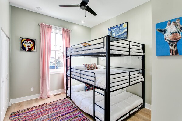 A bunk bed room with a ceiling fan, a window with pink curtains, and two paintings on the walls.