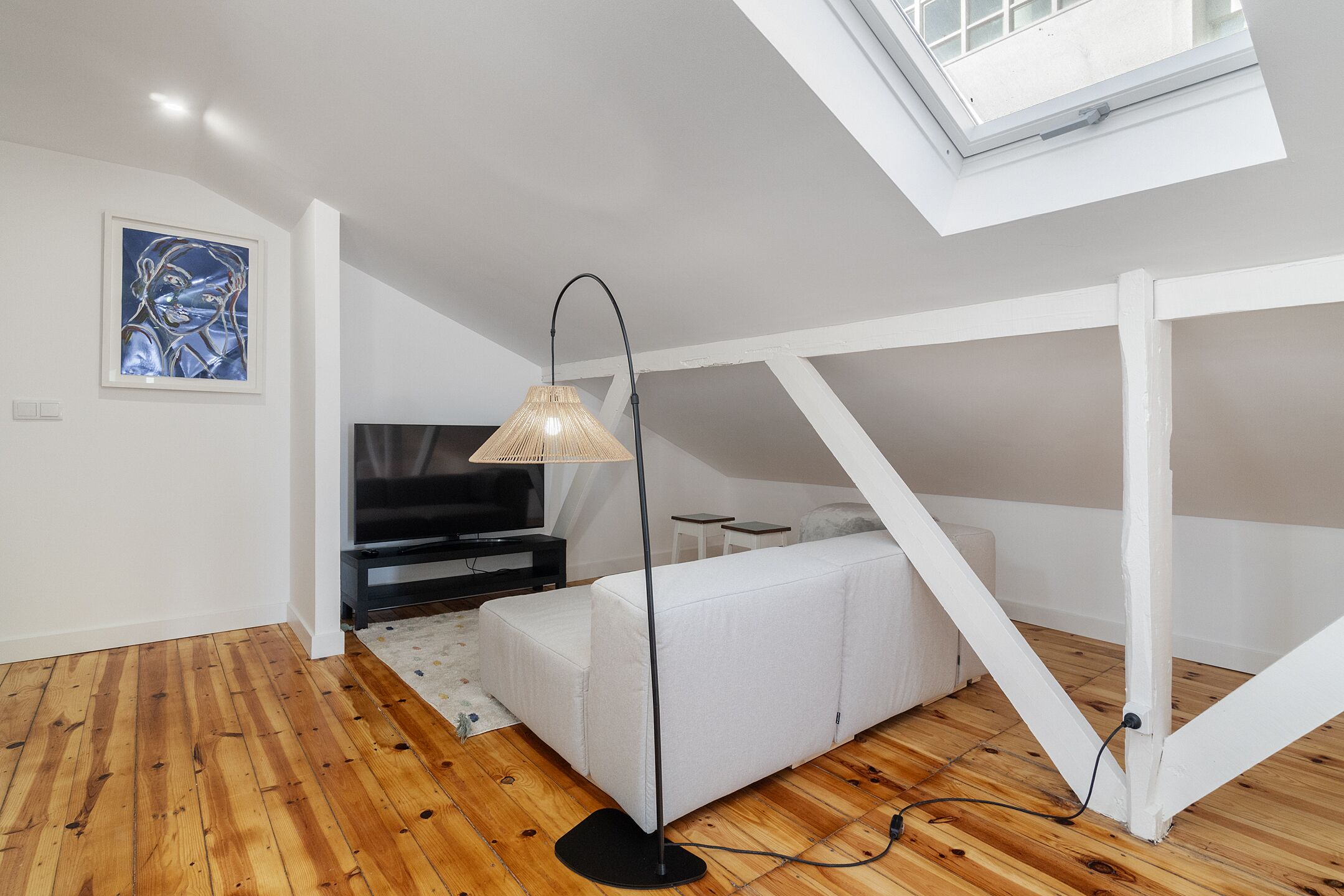 A modern loft bedroom with a sloped ceiling, a white sofa, a floor lamp, and a painting on the wall.