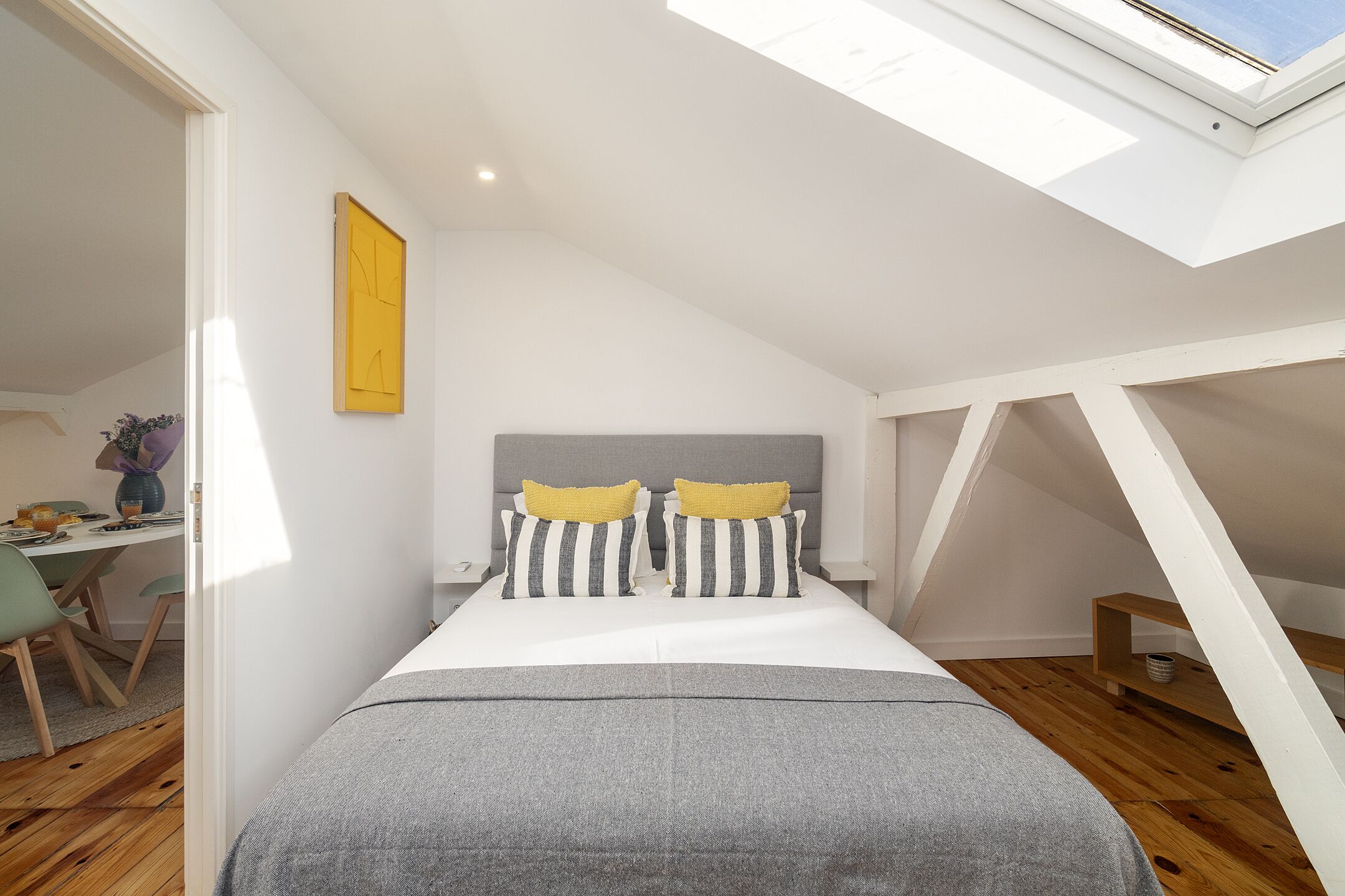 Quarto com claraboia, cama com almofadas listradas, escrivaninha com cadeira e piso de madeira.