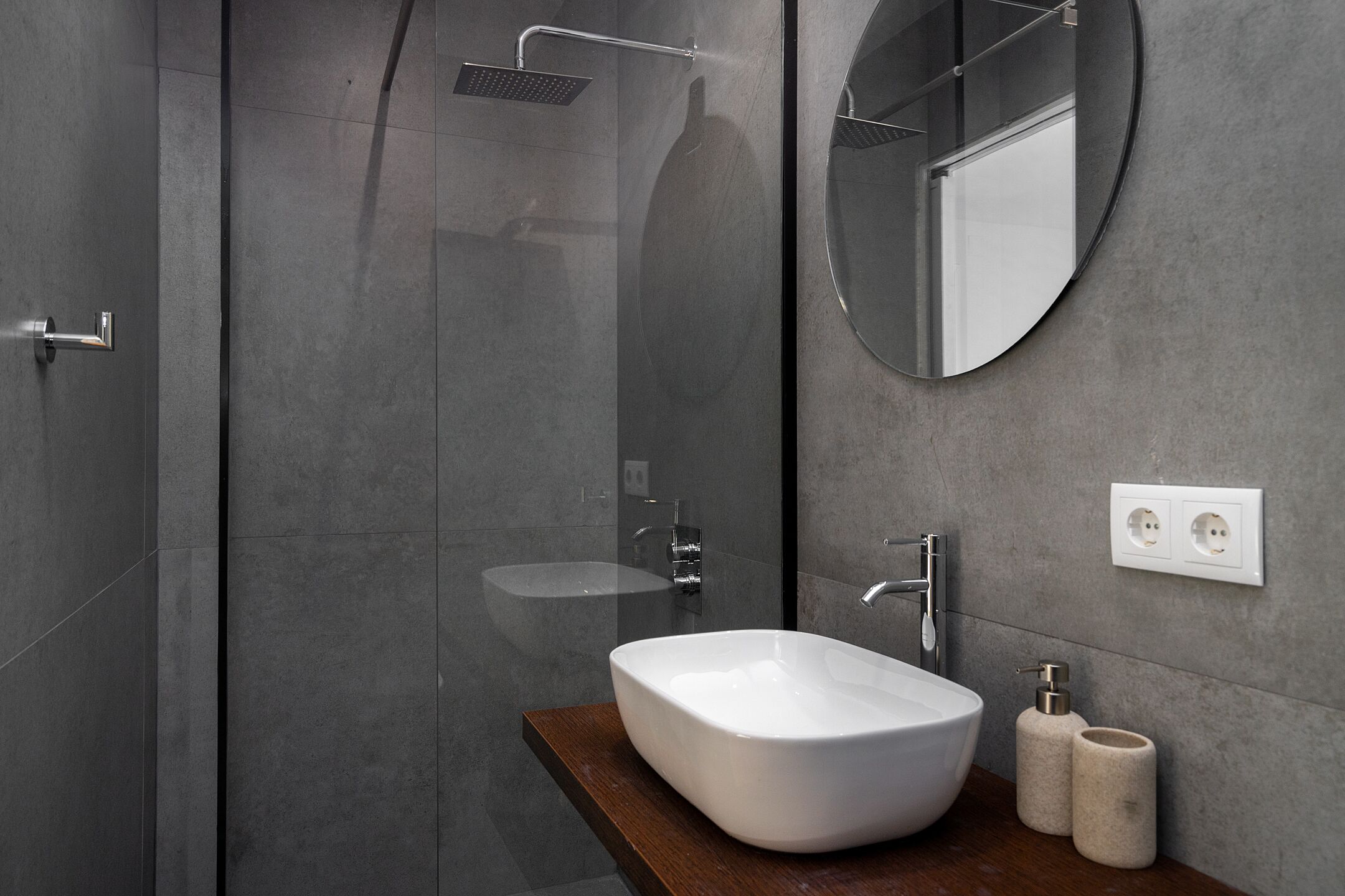 Een moderne badkamer met een douchecabine van glas, een witte wastafel en een ronde spiegel.