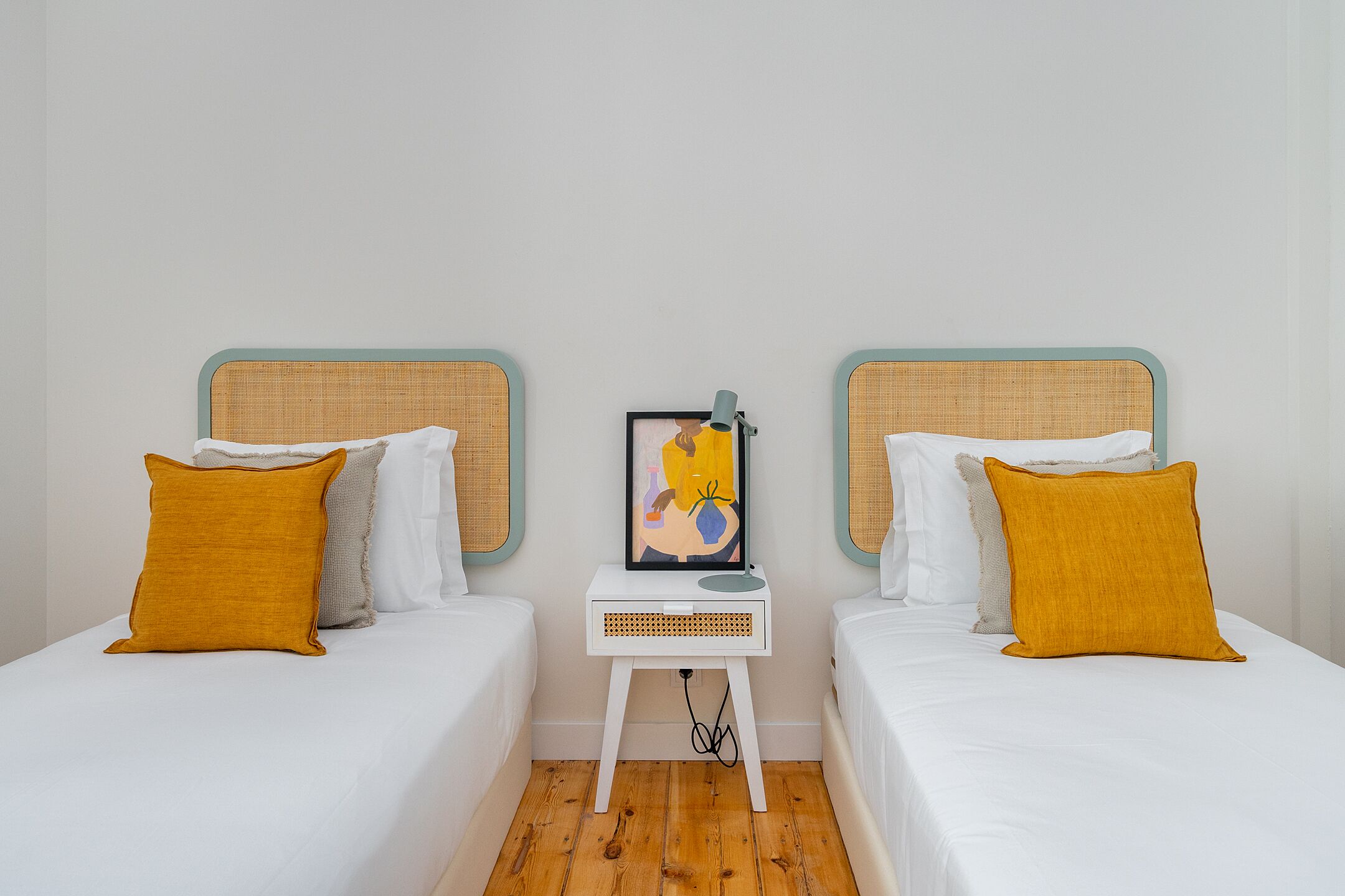 Una habitación con dos camas, cada una con colchón blanco y almohada color mostaza. Una mesita de noche pequeña, con un cuadro enmarcado, se encuentra entre las camas.