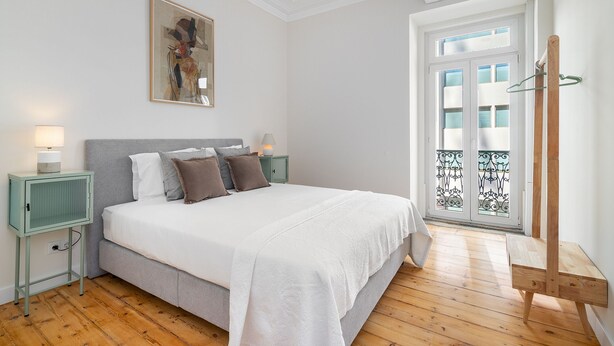 Quarto com cama, mesas de cabeceira, luminária pendente, janela com vista e porta para varanda.