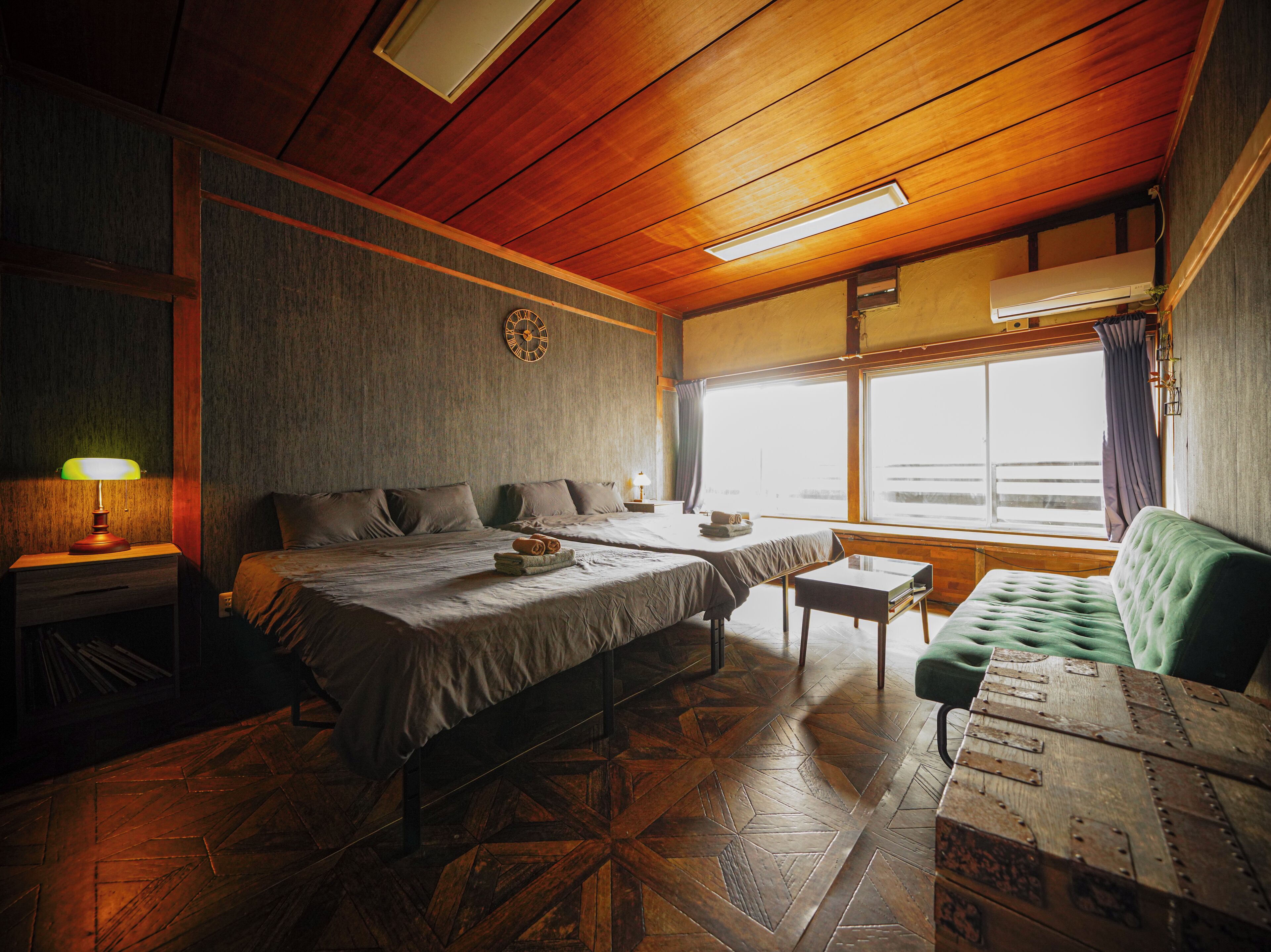 Una habitación con dos camas, una mesita pequeña, un sofá verde, un reloj en la pared y una ventana con cortinas.