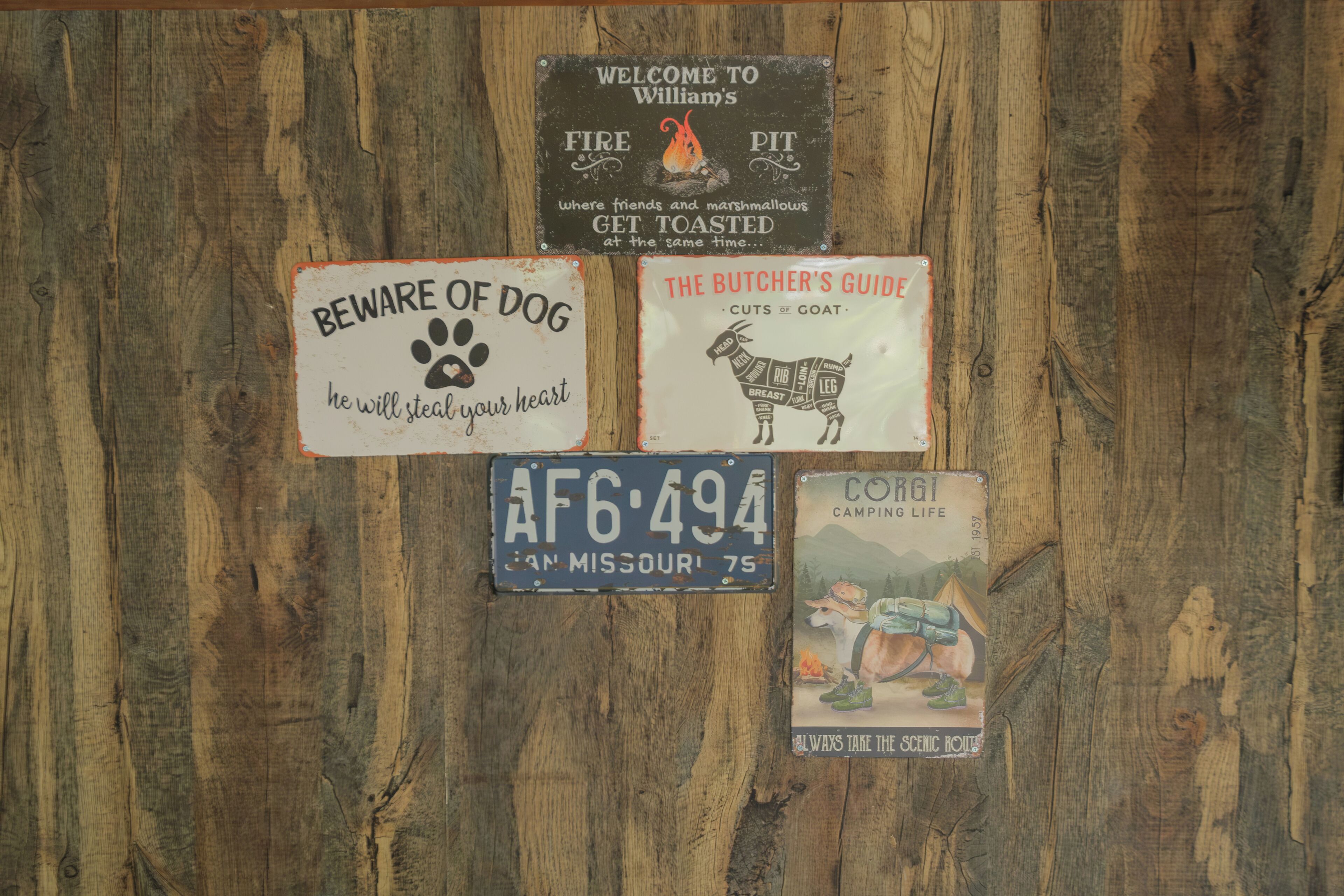 A wooden wall with various signs including a 'Beware of Dog' sign, a welcome sign, a butcher's guide, a license plate, and a camping life sign.