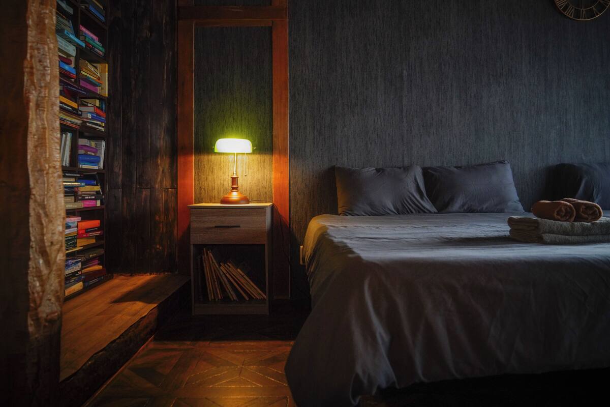 Un dormitorio con una estantería de madera, una mesita de noche con lámpara, una cama con almohadas y un reloj de pared.