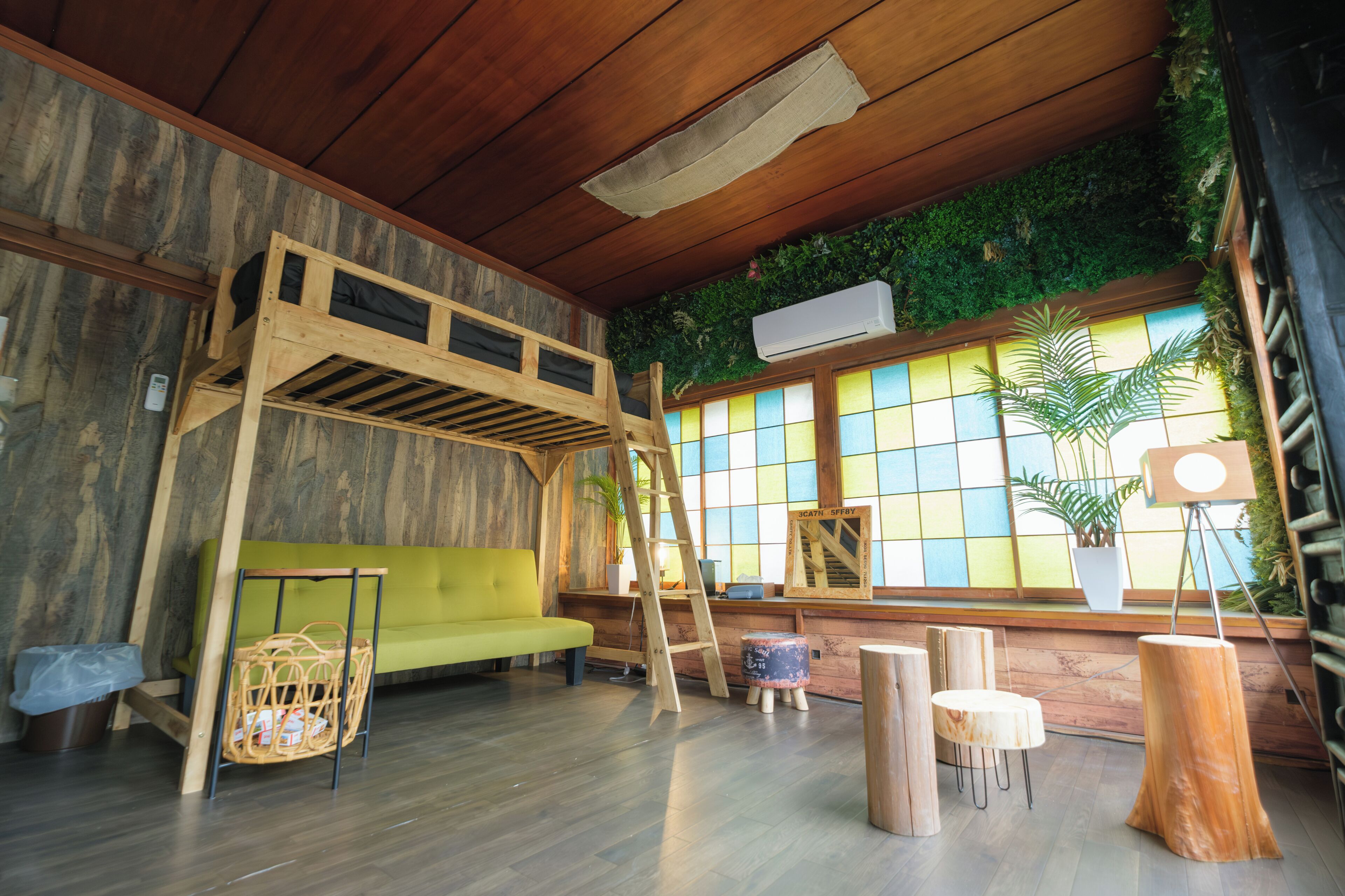Una habitación con una litera, un sofá verde, una escalera, una mesita y un amplio ventanal con vistas a la vegetación.