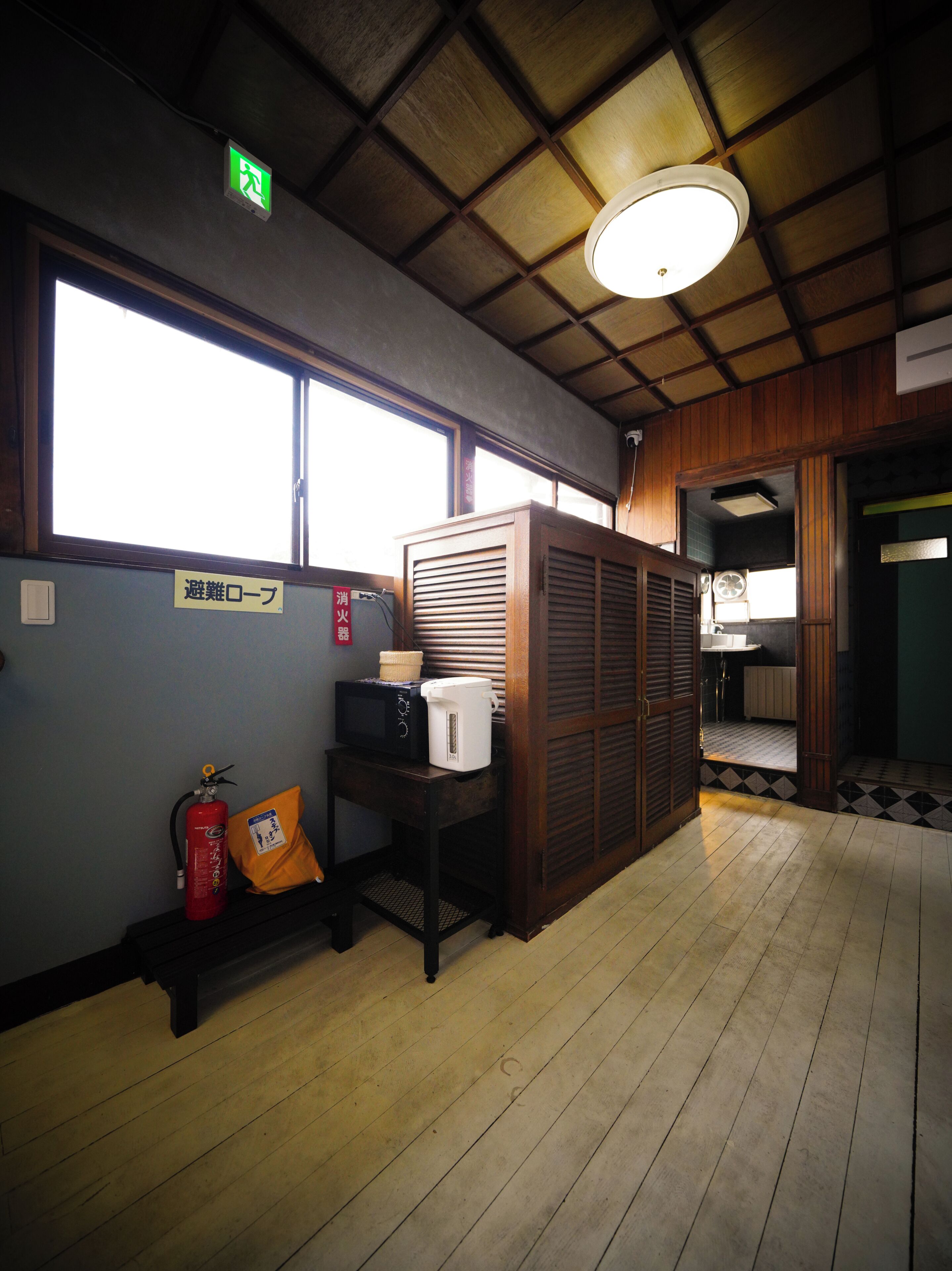 A room with a wooden ceiling, a window, a fire extinguisher, a table with a microwave and a kettle, and a door leading to another room.